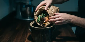 Close-up of hands depositing organic food scraps into a ceramic compost bin on a dark wooden kitchen counter, lit by a dramatic spotlight, illustrating waste reduction.