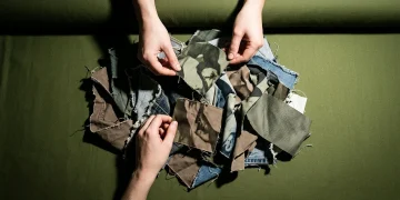 Overhead view of hands sifting through discarded textile scraps on an olive green fabric bolt, symbolizing hidden costs in fashion supply chains.